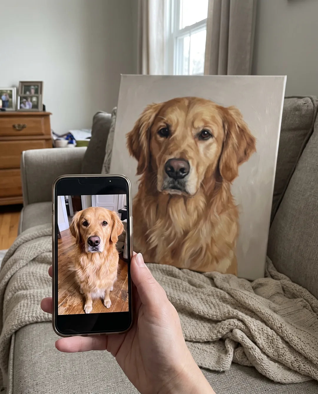 A phone held up showing the original dog photo next to the finished framed canvas portrait of the same dog, in a real living room