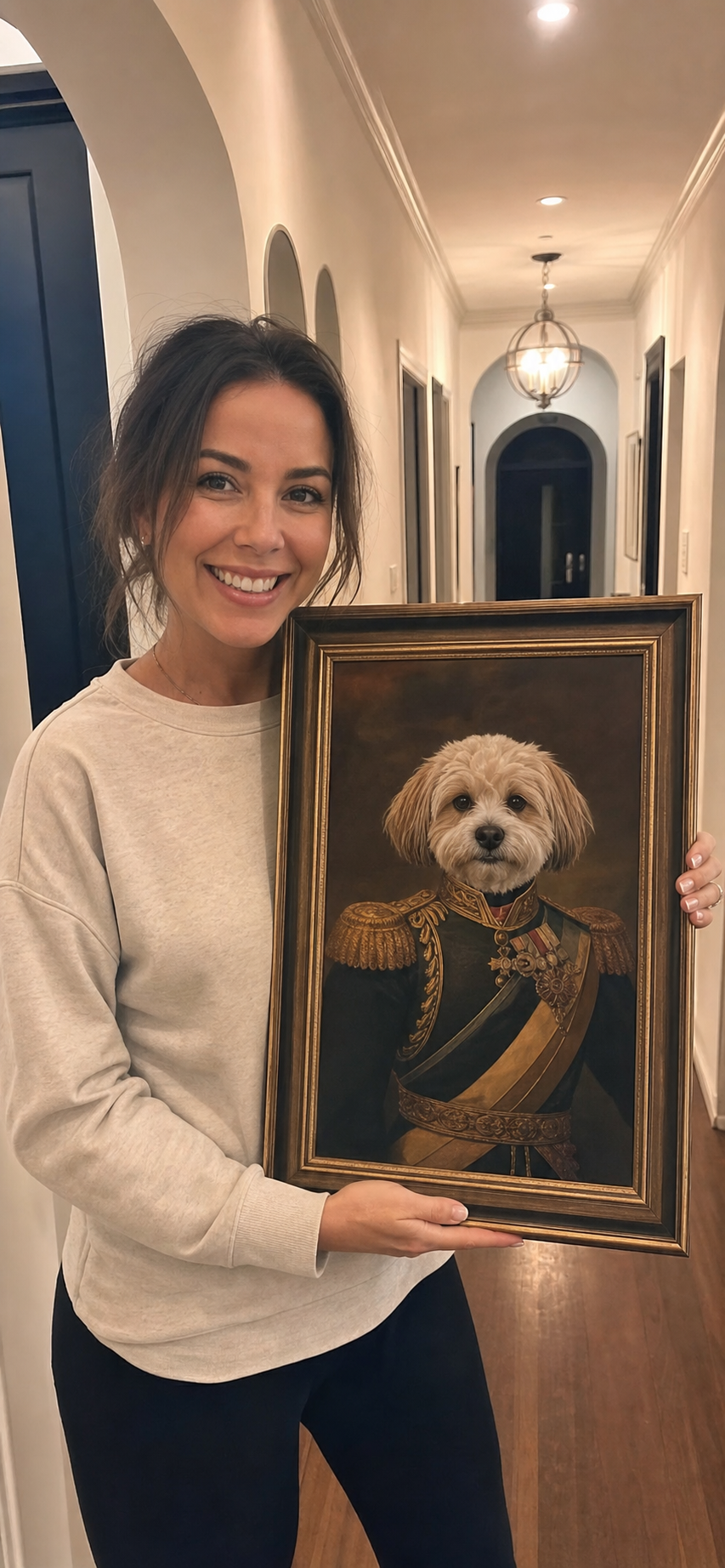 Smiling woman holding a framed pet portrait in a bright hallway at home
